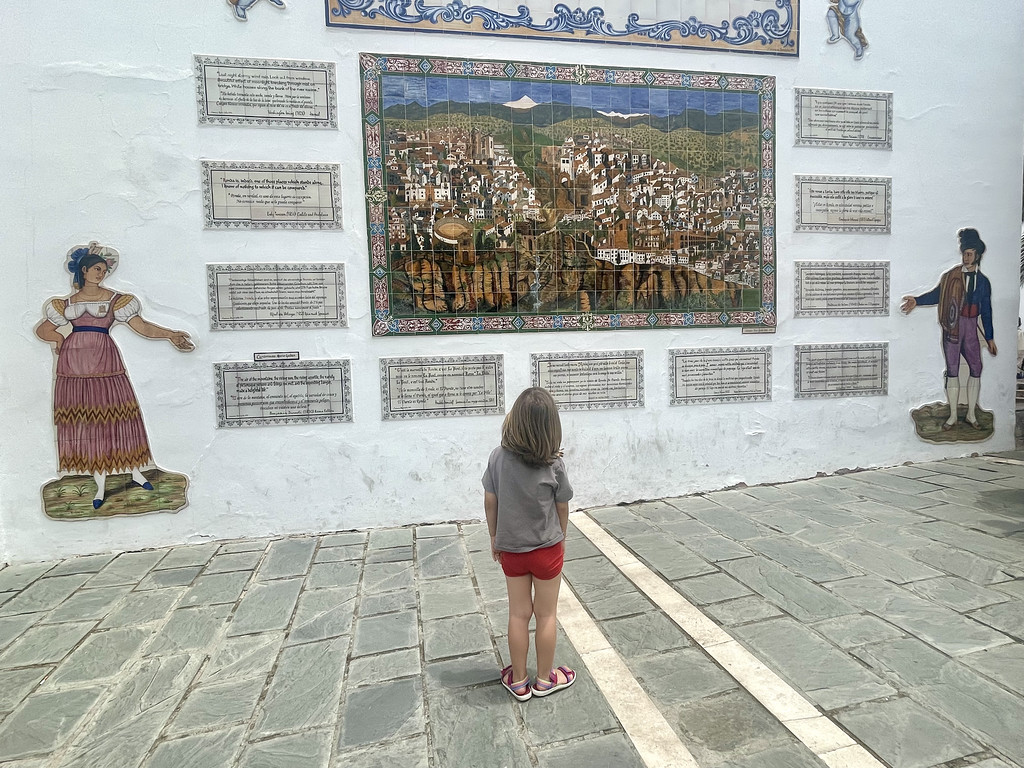 A child looking at a decorative tiled mural of Ronda on a whitewashed wall, flanked by painted costume cutouts.