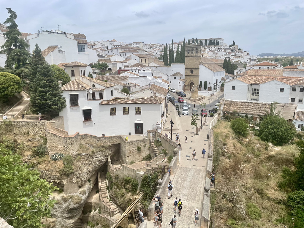 An elevated view of Ronda's white town buildings and a stone pathway leading toward an old gate, with tourists walking.