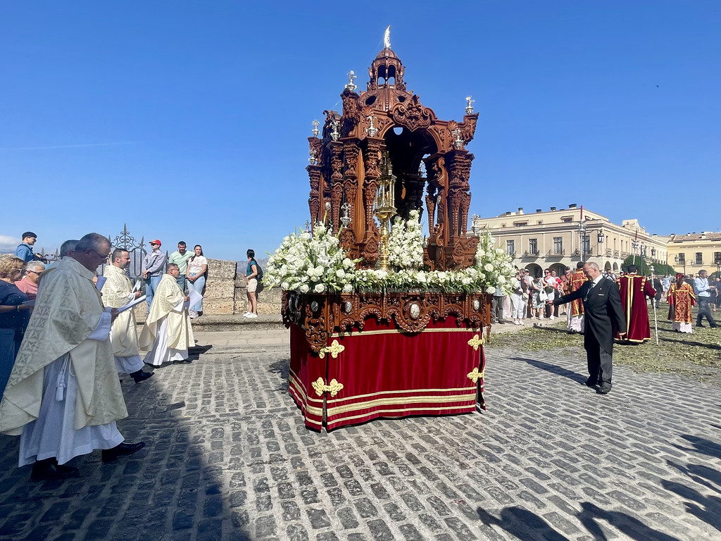 A religious Corpus Christi procession in Ronda with clergy in white and gold vestments beside an ornate flower-covered float.